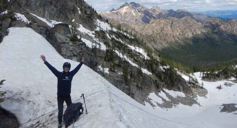 a person standing on a snowy bank in front of a mountainous landscape raises their hands in the air in celebration on a mountaineering course with outward bound